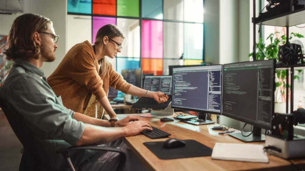 Male Programmer Shows Progress to Female Start-Up CEO on Desktop Computer in Creative Office Space. Caucasian Man and Woman Discussing New Features For Artificial Inteligence Software.