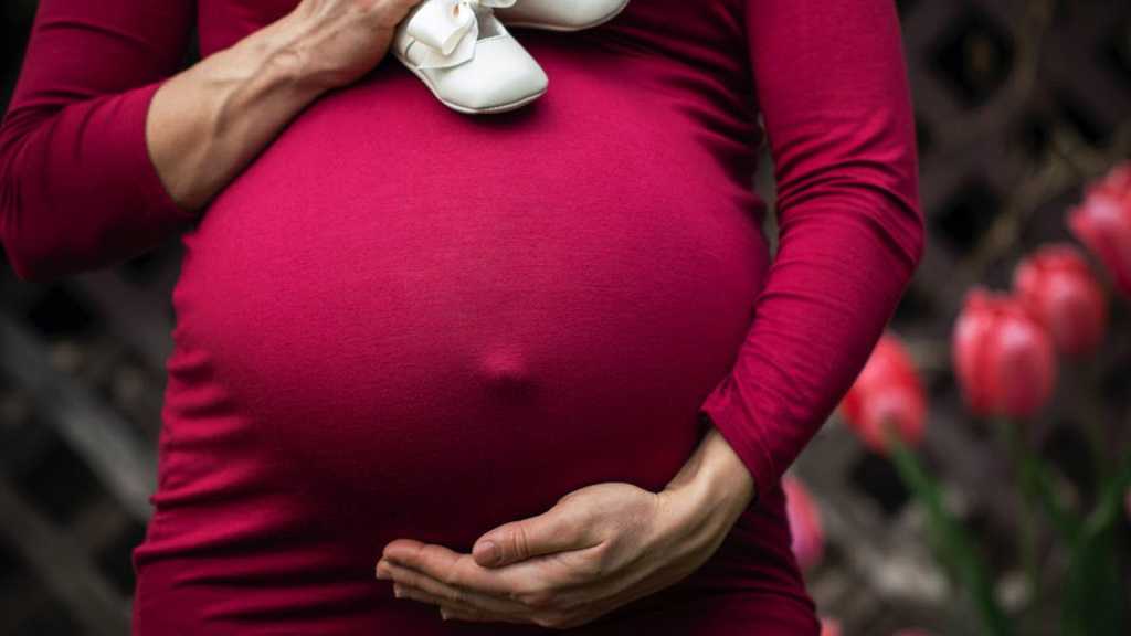 Pregnant woman in red dress holding belly with baby shoes