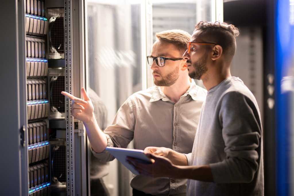 Team of young multi-ethnic specialists in glasses standing at server and using tablet while managing network server together in data center