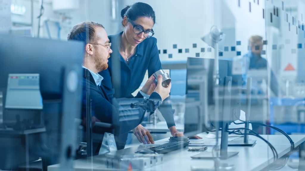 Modern Factory: Female Project Supervisor Talks to a Male Industrial Engineer who Works on Computer. They Inspect Machinery Mechanism and Design Improved Version. Working High-Tech Industrial Facility