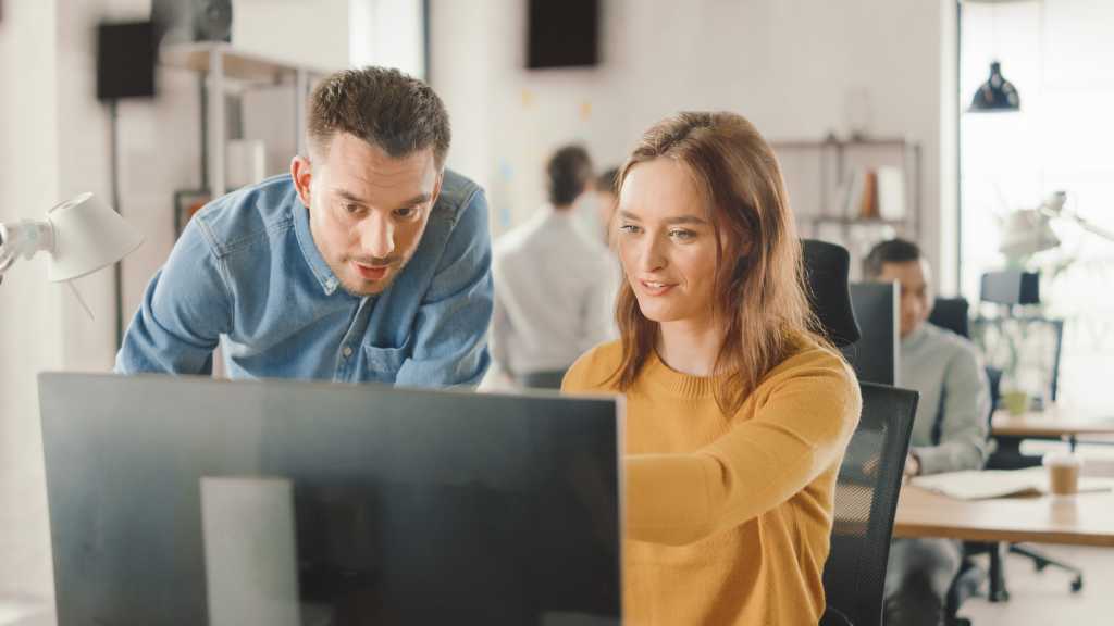 Female Specialist Works on Desktop Computer, Project Manager Stands Beside and gives Advice on Optimizing Workflow for Customer Experience Management. Modern Office with Diverse Team of Professionals