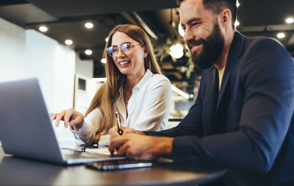 Cheerful businesspeople using a laptop in an office. Happy young entrepreneurs smiling while working together in a modern workspace. Two young businesspeople sitting together at a table.