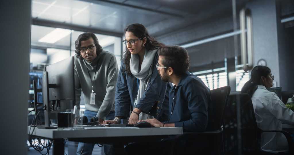 Team of Three Talented Young Software Engineers Use Computer to Discuss a Technological Project in Modern Industrial Office. Group of Male and Female Scientists Work in Research and Development Center