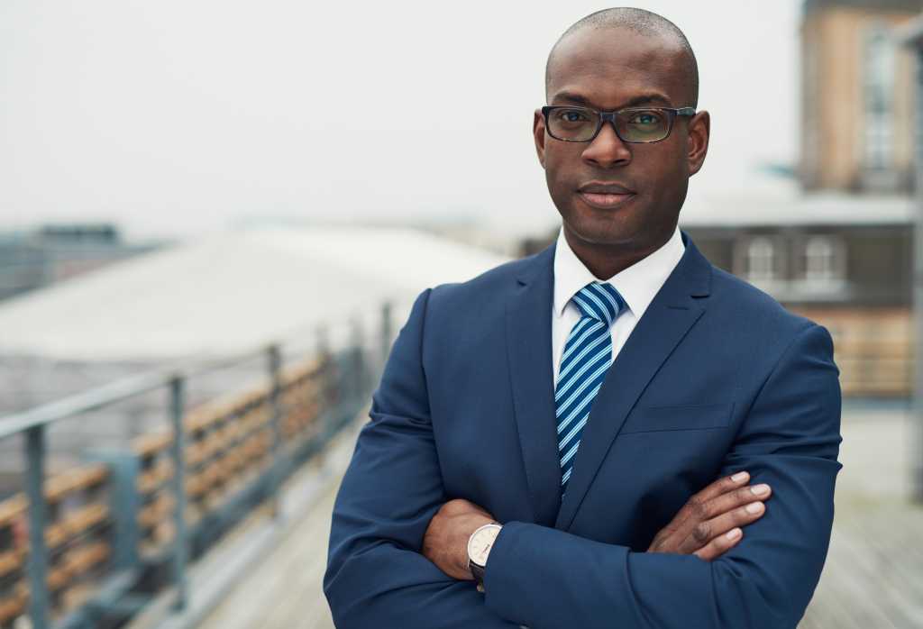 Confident black business man in a stylish suit standing with folded arms on a rooftop of n office block looking at the camera with a serious expression